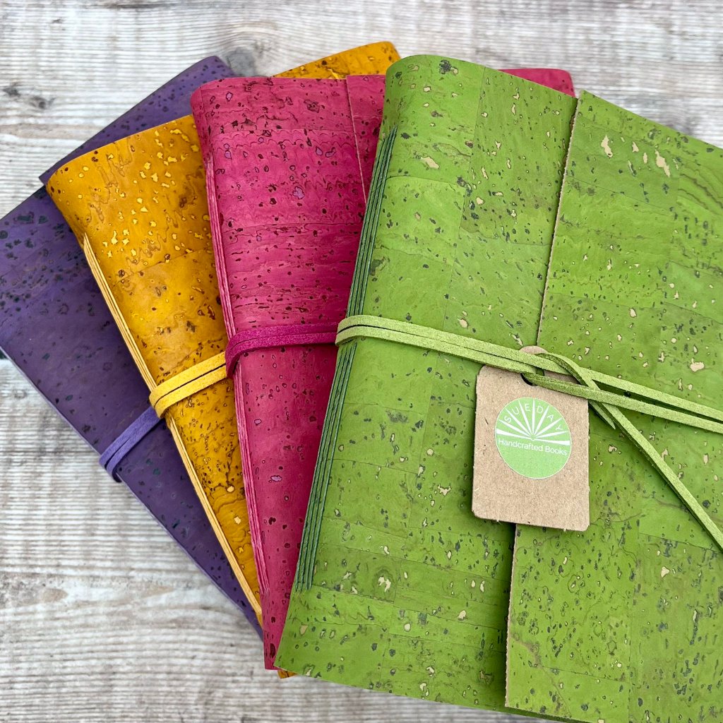 A stack of four vibrant, handcrafted journals with cork covers in shades of purple, yellow, magenta, and lime green, each tied with a coordinating strap and a small "Handcrafted Books" tag. The image is copyright Sue Day.