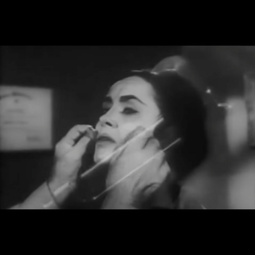 Screenshot still from The Wasp Woman (1959, horror film). In a black-and-white scene, a woman sits while another person’s hands pull and press at the skin on her cheeks as part of an experimental serum test. Used for commentary in a Hallowe’en body horror blog. Content note: the film contains scenes of animal testing/animal cruelty.